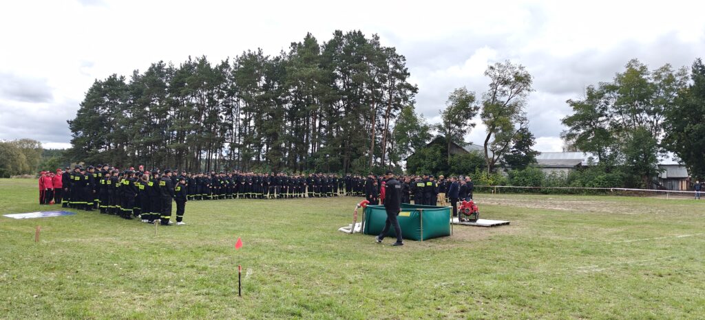 Grupa strażaków w czarnych mundurach z żółtymi napisami stoi na trawie. W tle drzewa i budynki. Po prawej zielony zbiornik z pompą.