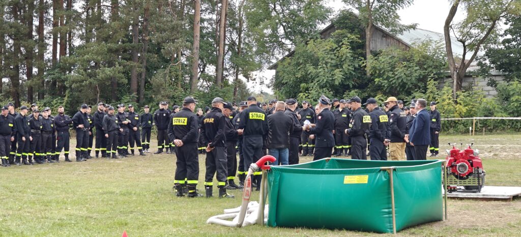 Grupa strażaków w czarnych mundurach zgromadzona na trawniku. W tle zielone drzewa i budynek. Na pierwszym planie zielony basen i sprzęt strażacki.