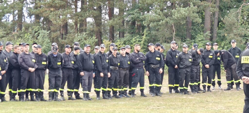 Na zdjęciu grupa osób w czarnych mundurach strażackich stoi na tle lasu. Mundury mają żółte elementy. W tle widoczne są drzewa iglaste.