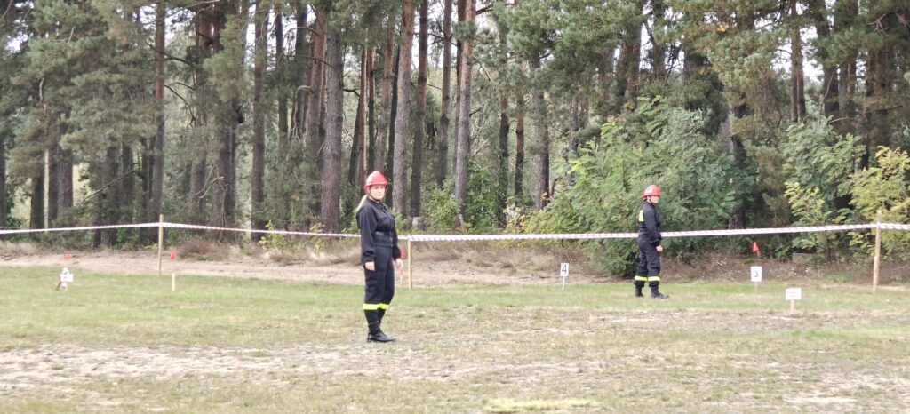 Na zdjęciu dwie osoby w czarnych mundurach strażackich i czerwonych kaskach na trawie. W tle las. Przed nimi taśma ostrzegawcza.