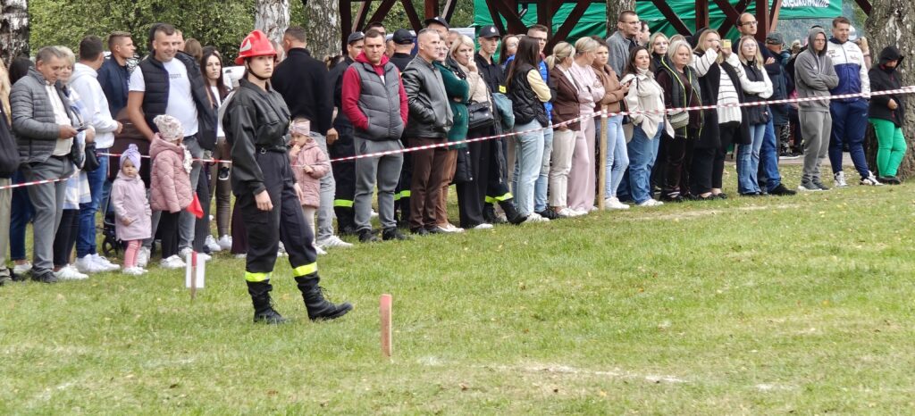 Ludzie stoją w grupie na trawie, odgrodzeni taśmą. Kobieta w stroju strażaka z czerwonym hełmem na pierwszym planie. W tle drzewa i zielony namiot.