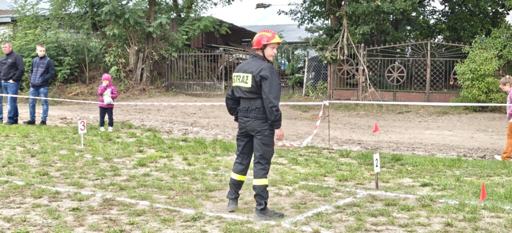 Mężczyzna w strażackim mundurze stoi na polu oznaczonym numerami. Tło to zieleń i ogrodzenie. Ludzie w kurtkach obserwują.