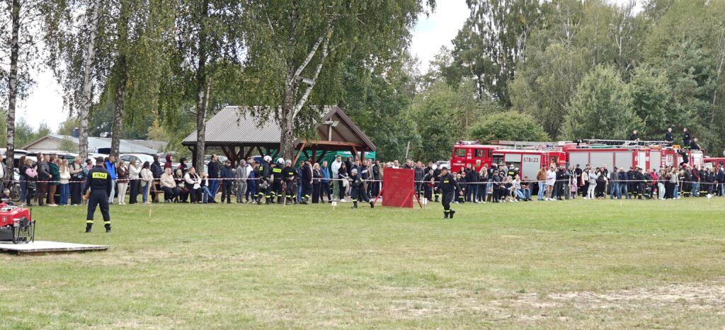 Strażacy w czarnych mundurach na zielonej trawie, za nimi tłum ludzi. W tle czerwone wozy strażackie i altana w parku.