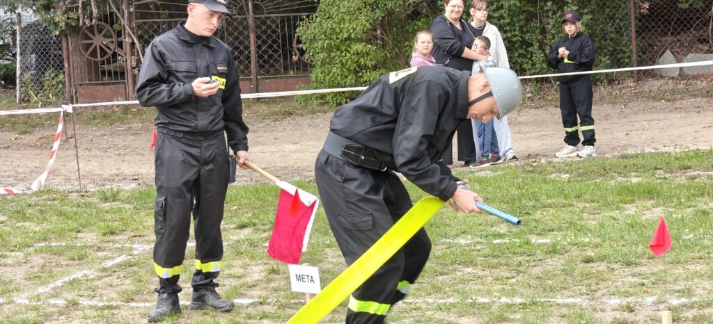 Dwóch mężczyzn w czarnych mundurach na trawie, przy czerwonej fladze i napisie meta. W tle ludzie i ogrodzenie. Kolory: zieleń, czerwień, czerń.