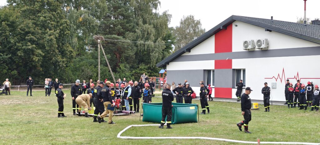 Grupa strażaków w czarnych i czerwonych kaskach ćwiczy na trawie przed biało-czerwonym budynkiem. W tle drzewa i widzowie. Atmosfera przypomina wydarzenie plenerowe.
