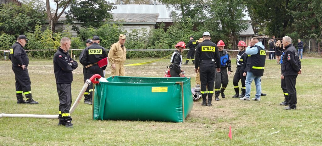 Grupa strażaków przy zielonym zbiorniku z wodą, na trawie. W tle drzewa i budynki. Ubrani w czarne mundury z odblaskowym napisem.