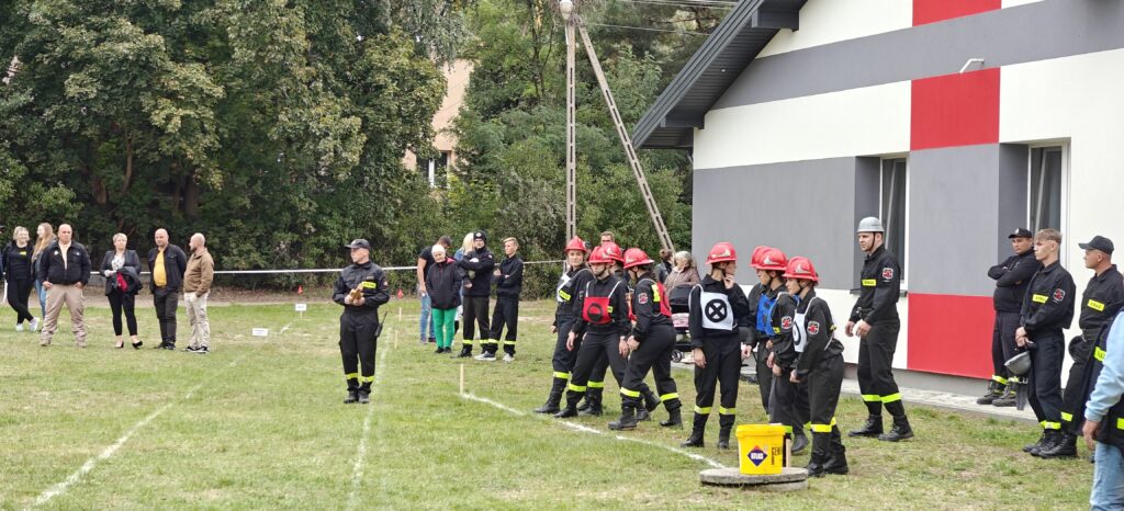 Grupa młodych strażaków w czerwonych kaskach stoi na trawie. Tło to szary budynek z czerwonymi akcentami i zielone drzewa. Obok znajdują się widzowie i inni strażacy.