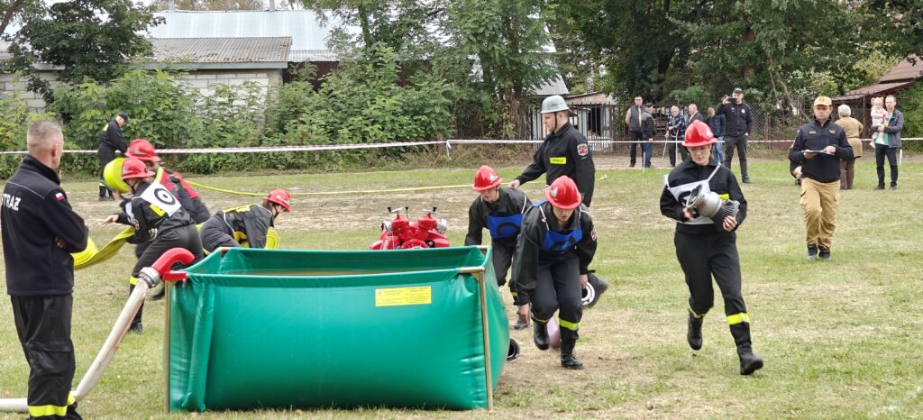 Strażacy w czerwonych kaskach biorą udział w ćwiczeniach na zielonej trawie. Zielony zbiornik na wodę oraz czerwony sprzęt strażacki. W tle drzewa i widzowie na tle budynków.
