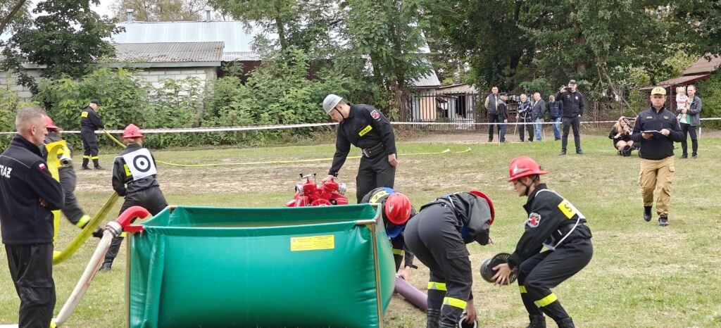 Na zdjęciu strażacy w czarnych mundurach i kaskach ćwiczą z zielonym zbiornikiem. W tle zieleń i budynki. Obserwatorzy stoją za taśmą ochronną.