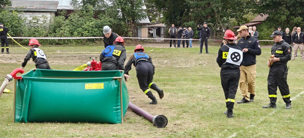 Zawody strażackie na zielonej łące, uczestnicy w kaskach i mundurach. Czerwony sprzęt strażacki obok zielonego zbiornika. Tło z drzewami i widzami.
