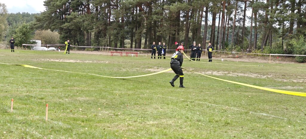 Osoba w strażackim mundurze biegnie z wężem przeciwpożarowym po trawiastym polu. W tle widać drzewa i grupę obserwatorów. Całość otoczona taśmą zabezpieczającą w kolorze biało-czerwonym.