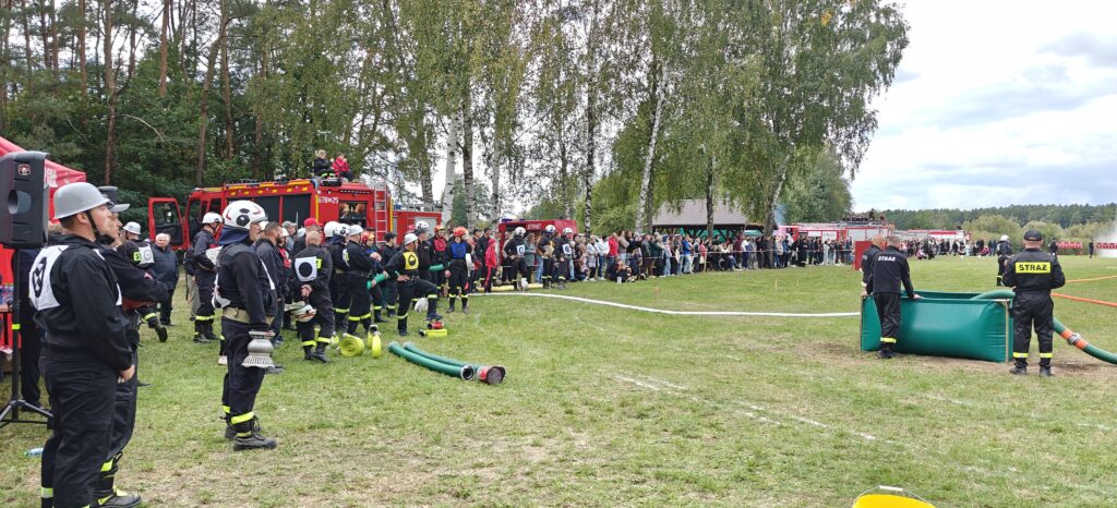 Strażacy w czarnych uniformach uczestniczą w ćwiczeniach na zielonej łące. W tle czerwone wozy strażackie i zebrany tłum. Obok drzewa i niebieskie niebo.