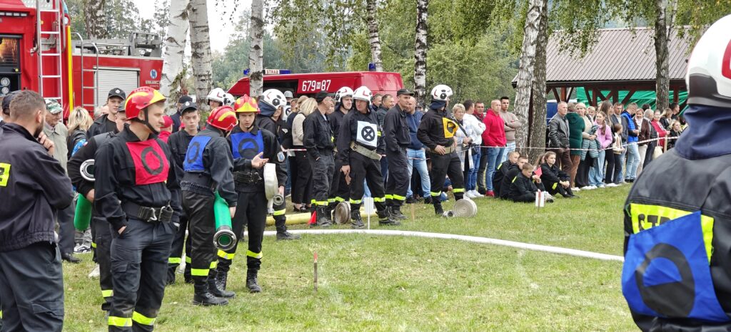 Grupa strażaków w hełmach i mundurach stoi na trawie z wozem strażackim w tle. Tło tworzą drzewa i zbierająca się publiczność. Obraz jest pełen zieleni i czerwieni.