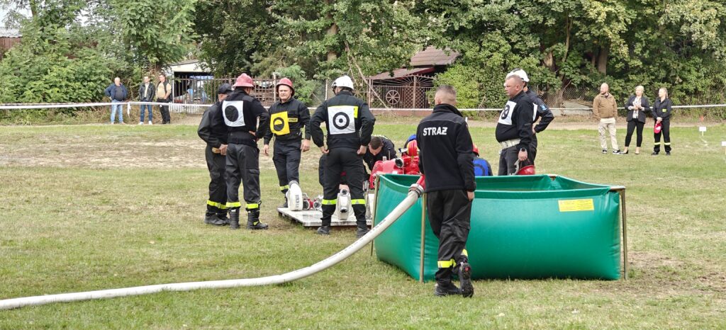Strażacy w hełmach przygotowują sprzęt na zielonym trawniku. Tło to drzewa i kilku obserwatorów. Widoczny duży, zielony pojemnik.