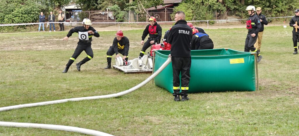 Strażacy w czarnych mundurach uczestniczą w zawodach na trawie. Zielony zbiornik z wodą na pierwszym planie. W tle drzewa i ogrodzenie.