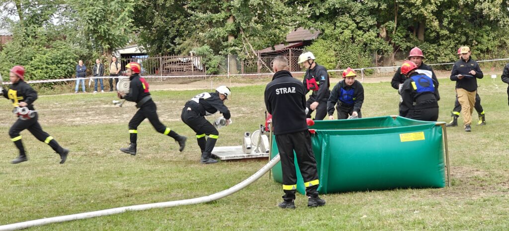 Strażacy w czarnych mundurach i kolorowych hełmach ćwiczą na zielonej trawie. Obok stoi zielony zbiornik wodny. W tle drzewa i widzowie.