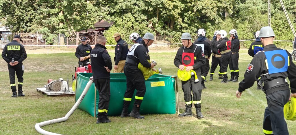 Strażacy w czarnych mundurach i kaskach pracują przy zielonym zbiorniku z wodą. Na trawie stoją węże strażackie. W tle widać drzewa i ogrodzenie.