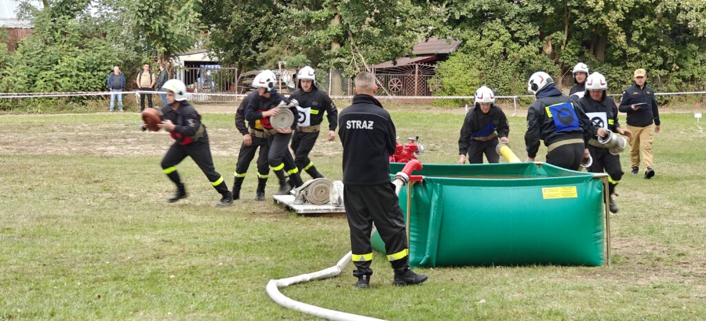 Strażacy w ciemnych mundurach ćwiczą z wężami pożarniczymi na trawie. Tło pokrywają drzewa i budynki. Na środku stoi zielony zbiornik wody.