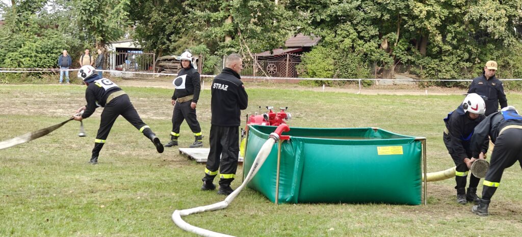 Strażacy w czarnych mundurach ćwiczą z wężami wodnymi na trawie. Na środku zielony zbiornik i czerwone urządzenie. W tle drzewa i ogrodzenie.