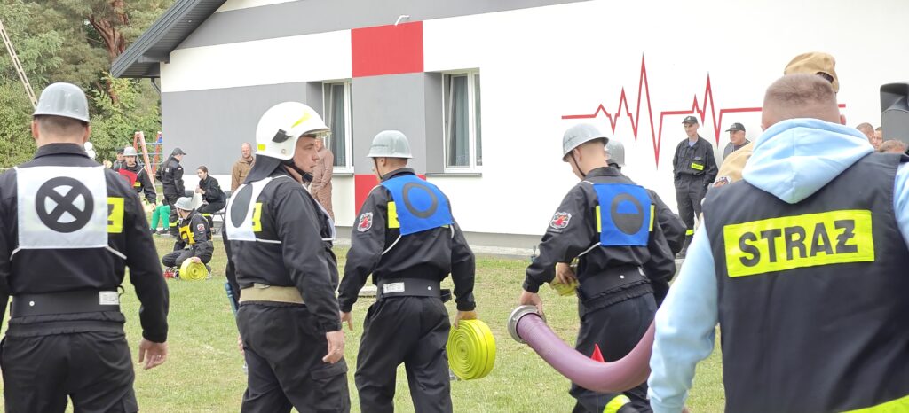 Strażacy na zielonej trawie przy szarej budowie, trzymają węże. Ubrani w czarne mundury, z napisami STRAŻ na plecach. W tle biały budynek z czerwonym wzorem.