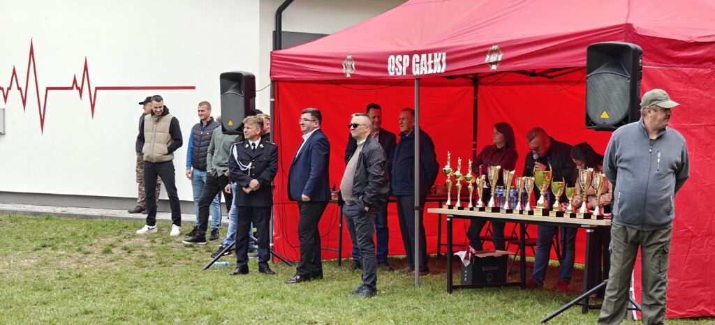Ludzie stoją pod czerwonym namiotem z trofeami na stole. Tło to biała ściana z czerwoną falą w kształcie serca. Trawnik na pierwszym planie.
