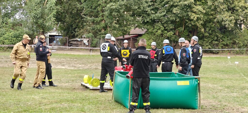 Grupa strażaków w hełmach obok zielonego zbiornika podczas ćwiczeń. Tło to drzewa i trawa. Czarne i żółte uniformy.