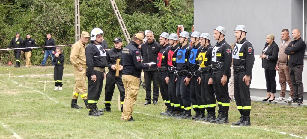 Strażacy w mundurach stoją w rzędzie. Obok, ludzie w ciemnych ubraniach obserwują. Tło to zielone krzewy i szara ściana.