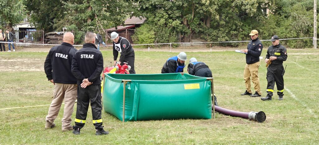 Ćwiczenia straży pożarnej na trawie, zielony zbiornik, czerwone pompy. W tle drzewa i drewniany płot.