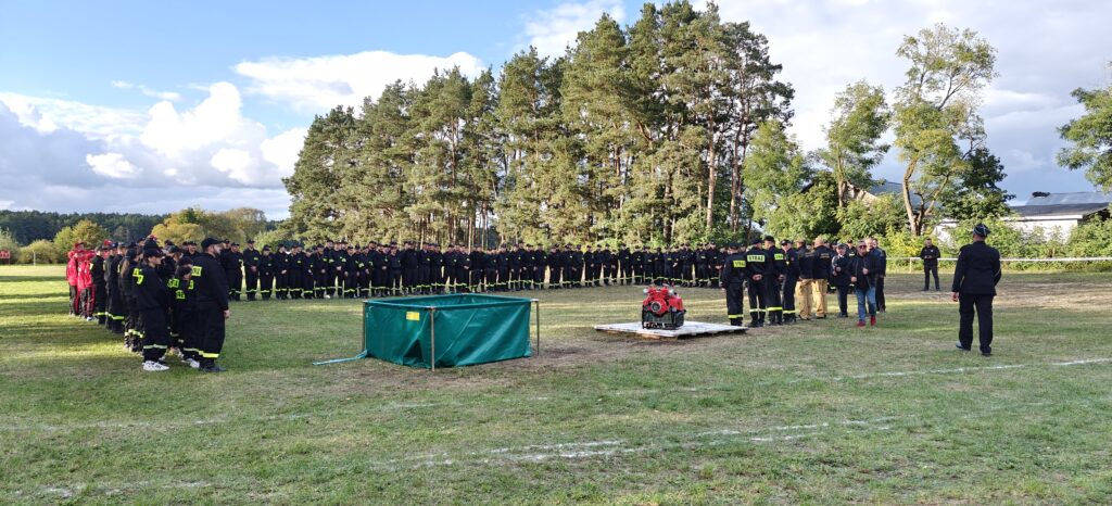 Grupa strażaków stoi na zielonej trawie, na tle drzew. Ubrani w czarne mundury z żółtymi elementami. Na ziemi widoczna czerwona motopompa.
