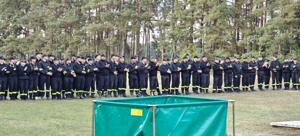 Grupa osób w czarnych mundurach stoi na tle lasu. Mają odblaskowe paski na spodniach. Z przodu zielone ogrodzenie.