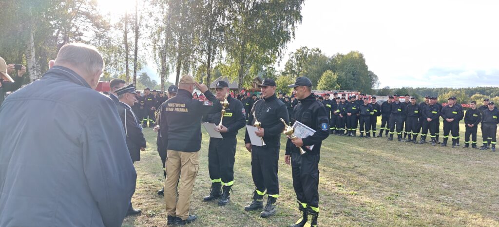 Grupa strażaków w czarnych mundurach ustawia się na trawie, w tle drzewa i niebo. Niektórzy trzymają puchary i dokumenty. Słońce świeci zza drzew.