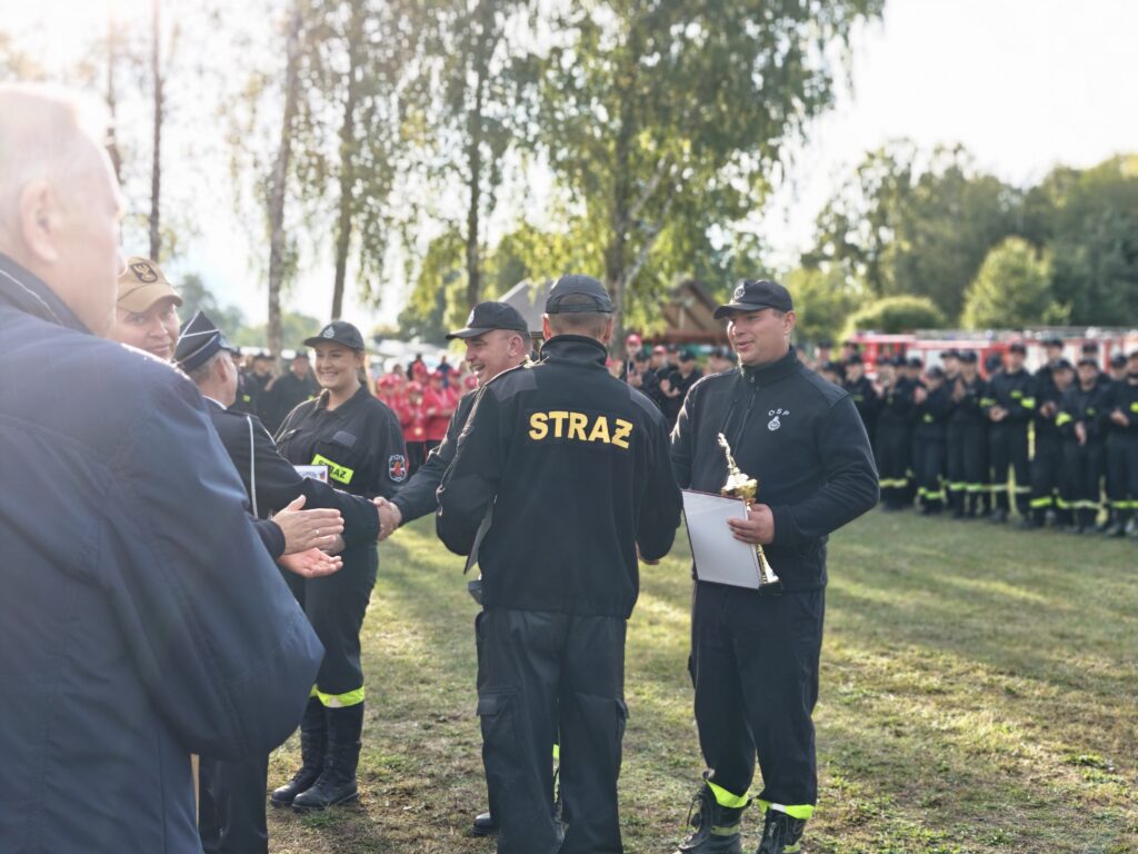 Grupa strażaków w ciemnych mundurach uczestniczy w ceremonii. Jedna osoba trzyma puchar i dokument. W tle zielone drzewa.