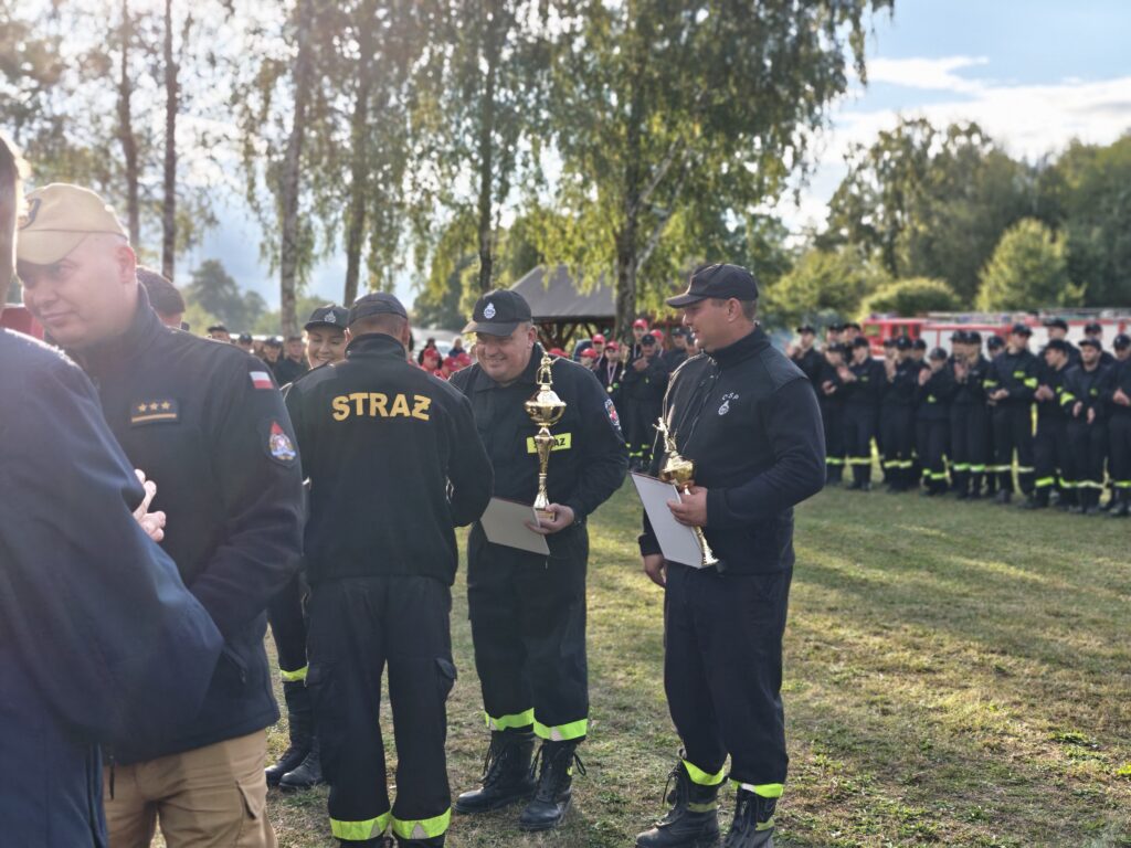 Strażacy w czarnych mundurach z napisami „STRAŻ” trzymają puchary na tle zieleni i błękitnego nieba. W tle tłum ludzi w podobnych uniformach. Drzewa dodają naturalnego uroku scenie.