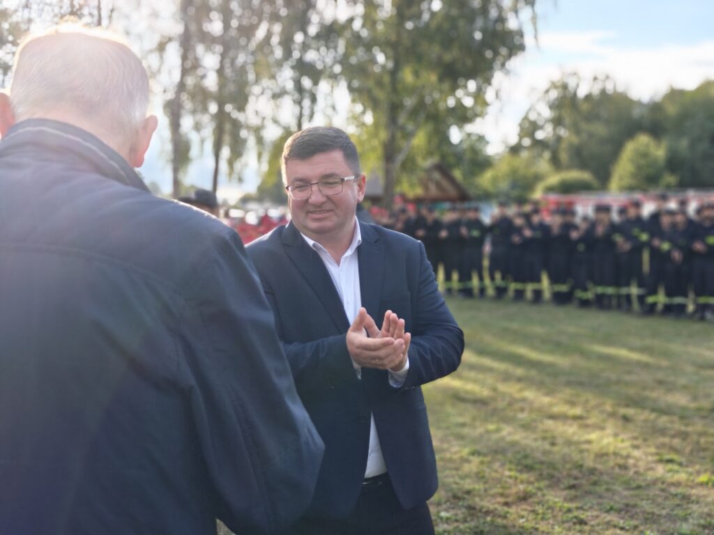 Mężczyzna w garniturze stoi na trawie i bije brawo. W tle grupa osób w ciemnych uniformach. Dookoła zielone drzewa.