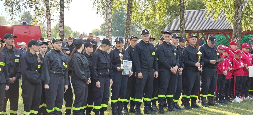 Grupa strażaków w czarnych mundurach stoi na trawie, z trofeum i dyplomem. Obok dzieci w czerwonych strojach sportowych. W tle drzewa i budynek.