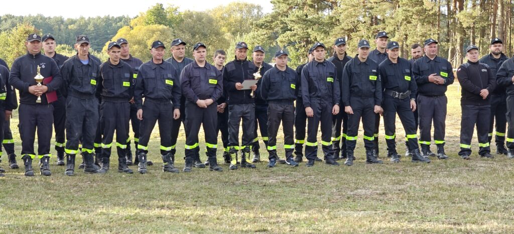 Grupa strażaków w czarnych mundurach stoi na trawie. W tle zielony las. Dwóch trzyma puchar i dyplom.