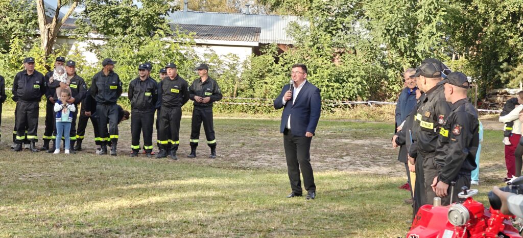 Grupa mężczyzn w ciemnych mundurach stoi na trawie, są różne ocienia zieleni. Osoba w garniturze mówi przez mikrofon. Tło z drzewami i budynkiem.