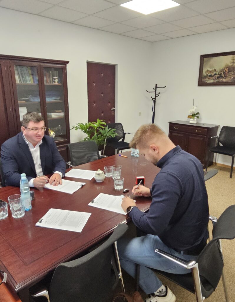 Dwóch mężczyzn przy biurku z dokumentami, ściany białe, obraz i regał w tle, drewniany stół i krzesła dookoła.