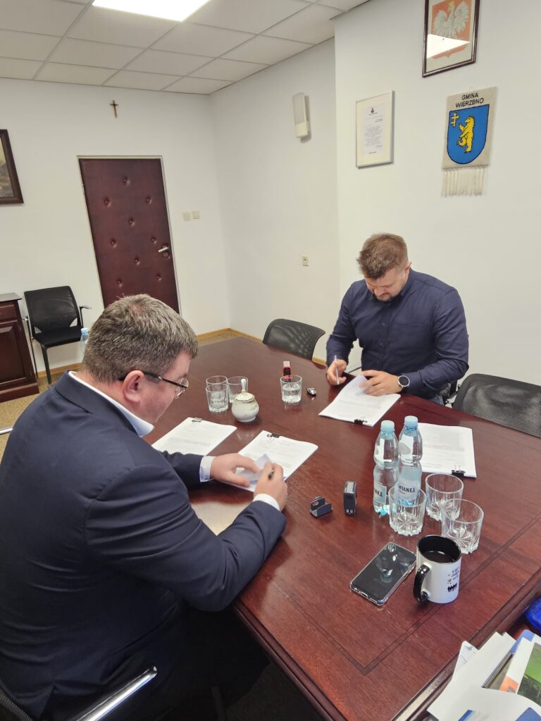 Dwóch mężczyzn w garniturach siedzi przy drewnianym stole, przeglądając dokumenty. Na stole są butelki wody, filiżanka i telefon. W tle biała ściana z herbem i krzyżem.