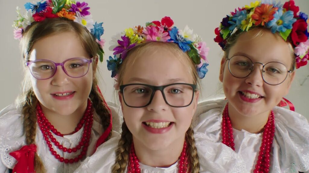 Trzy dziewczynki w okularach, ubrane w białe stroje ludowe. Na głowach mają kolorowe wianki z kwiatami. Tło jest jasne i neutralne.