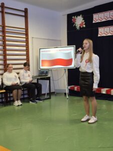 Na zdjęciu znajduje się uczennica śpiewająca podczas obchodów Święta Niepodległości. Ma na sobie białą bluzkę i czarną spódnicę, stoi na zielonej podłodze. W tle jest ekran z flagą Polski i napisami, a po lewej siedzi