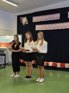 Trzy osoby stoją na scenie, trzymają kartki, ubrani na biało-czarno z kokardami. Tło ciemne z dekoracjami w kolorach biało-czerwonych.