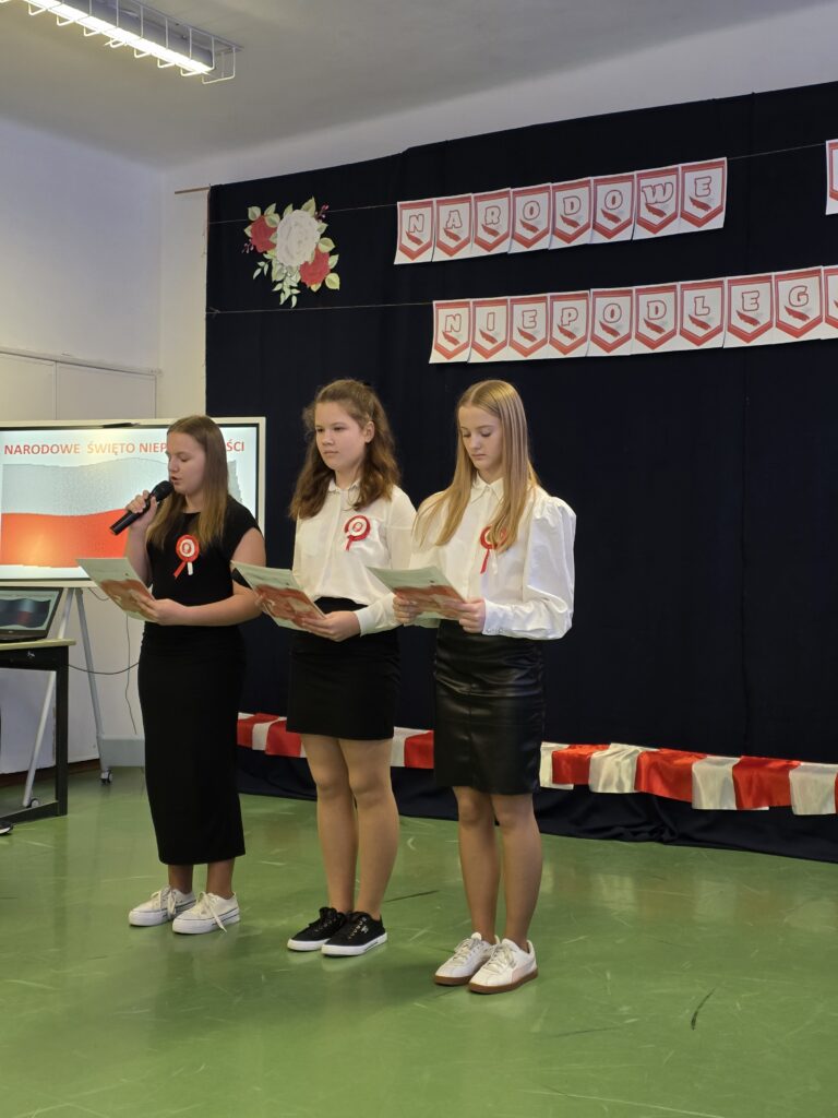 Trzy osoby stoją na scenie, trzymają kartki, ubrani na biało-czarno z kokardami. Tło ciemne z dekoracjami w kolorach biało-czerwonych.