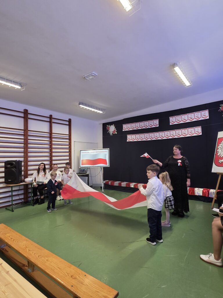 Dzieci trzymają flagę biało-czerwoną na zielonej podłodze. W tle tablica z napisem Narodowe Święto Niepodległości oraz dekoracje w barwach polskich. Po prawej stronie stoi kobieta w czarnej sukience.