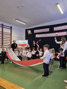 Dzieci w sali szkolnej trzymają biało-czerwoną flagę. Ubrane są w białe bluzki, tło ozdobione jest plakatami nawiązującymi do święta. Na ścianach widać dekoracje w narodowych kolorach.