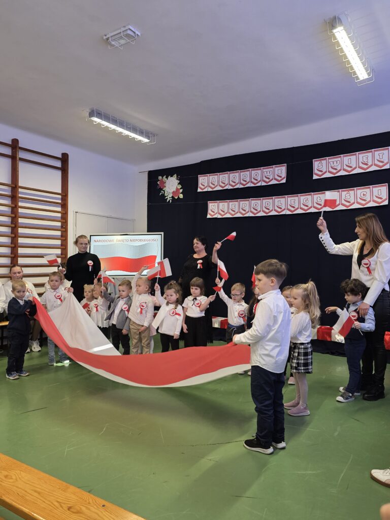 Dzieci w sali szkolnej trzymają biało-czerwoną flagę. Ubrane są w białe bluzki, tło ozdobione jest plakatami nawiązującymi do święta. Na ścianach widać dekoracje w narodowych kolorach.