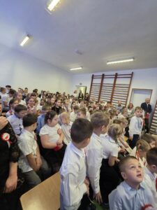 Sala pełna dzieci w białych koszulach, siedzących na krzesłach. Jasne oświetlenie i drabinki na ścianie. Uczniowie skupieni, część nosi kokardy.