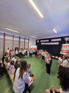 Dzieci ubrane na biało-czerwono w kręgu, trzymają polskie flagi. Zielona podłoga, drewniane ławki po lewej, ciemne tło z symboliką narodową.