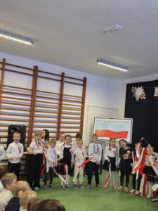 Dzieci w odświętnych strojach występują z flagami Polski. Tło jasne z brązowymi drabinkami i ekranem. Podłoga zielona.