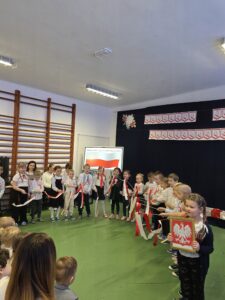 Grupa dzieci w białych koszulach z czerwonymi kokardami stoi na tle ciemnej ściany z polskimi flagami. Dzieci trzymają biało-czerwone wstążki, jedna z nich trzyma herb Polski. Tło jest ozdobione plakatami o Święcie